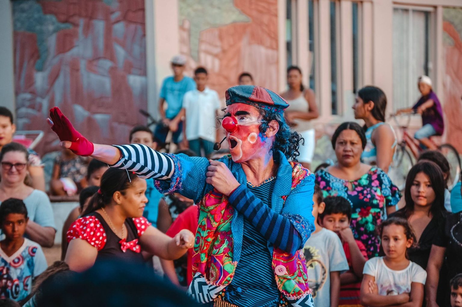 Artista caracterizado de palhaço se apresenta na rua diante de um público diverso.